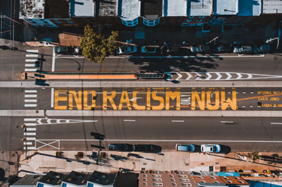 Abgebildet ist eine Straße in New York aus der Vogelperspektive. Mit orangenen Buchstaben ist auf Englisch geschrieben, dass der Rassismus beendet werden soll.